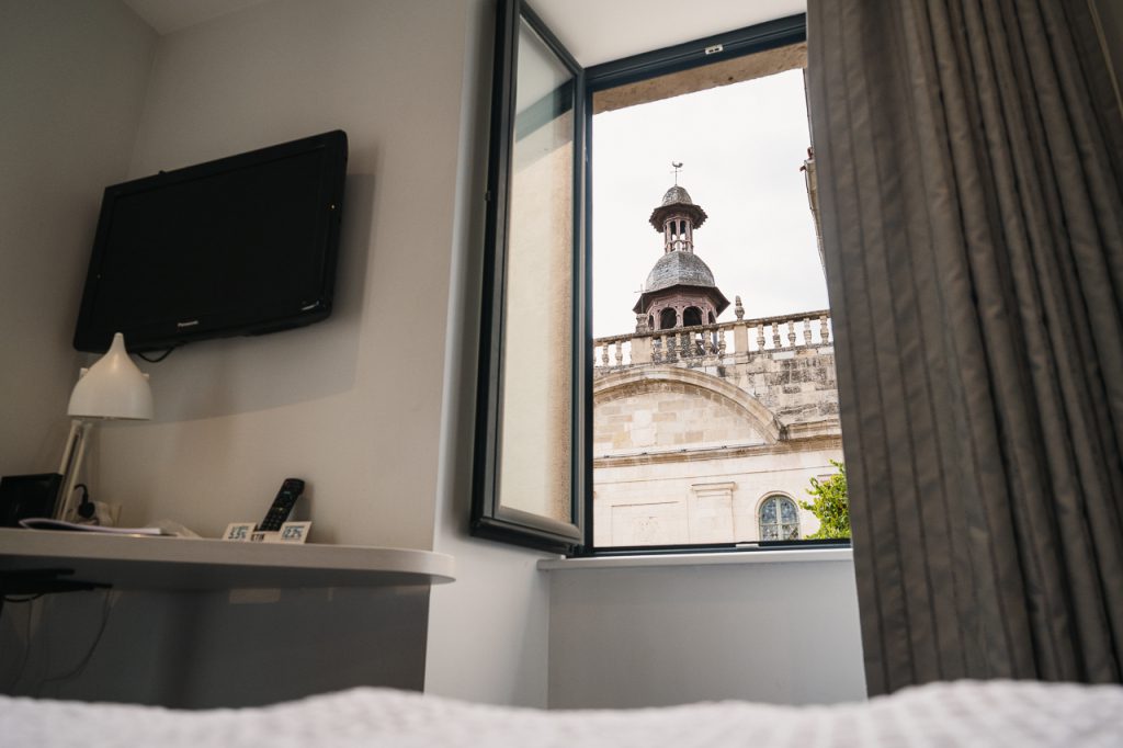 Chambre lumineuse de l'Hôtel Les Fleurines à Villefranche-de-Rouergue, avec un bureau fonctionnel et une fenêtre ouverte offrant une vue imprenable sur la chapelle des Pénitents Noirs. Un cadre paisible et inspirant pour travailler ou se détendre, en plein cœur de l'Aveyron.