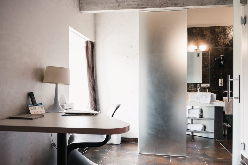 Chambre spacieuse et lumineuse de l'Hôtel Les Fleurines à Villefranche-de-Rouergue, équipée d'un bureau fonctionnel. À l'arrière, une salle de bain moderne avec une porte vitrée, offrant un design épuré et une ambiance agréable. Un cadre parfait pour un séjour alliant confort et style, au cœur de l'Aveyron.
