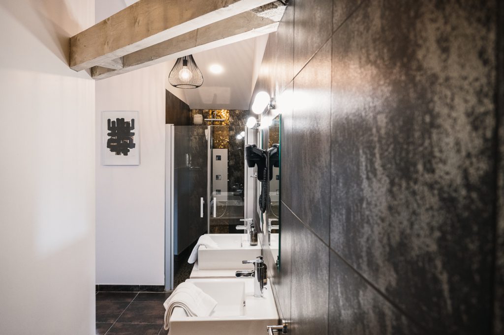 Salle de bain raffinée de l'Hôtel Les Fleurines à Villefranche-de-Rouergue, avec une crédence dorée, un carrelage foncé, et une touche artistique grâce à une affiche de Pierre Soulages sur le mur. Cette combinaison d'éléments modernes et d'art contemporain crée un espace élégant et apaisant pour se ressourcer en plein cœur de l'Aveyron.