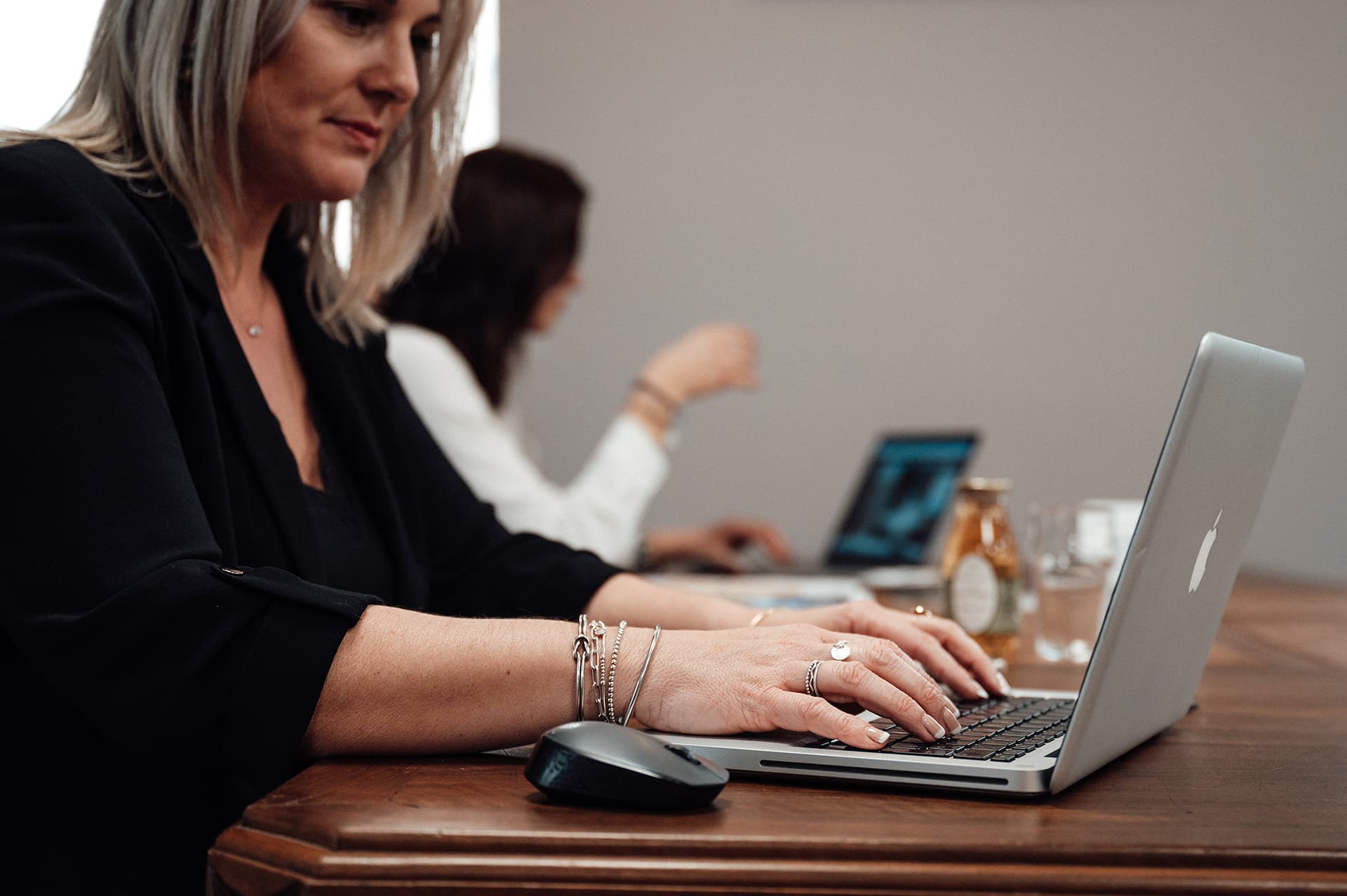 Deux femmes travaillant dans l’espace de coworking élégant et chaleureux de l’Hôtel Les Fleurines à Villefranche-de-Rouergue, Aveyron. Au premier plan, une femme en veste noire utilise un MacBook avec une souris posée sur une table en bois, portant des bracelets et une bague argentée. En arrière-plan, une autre femme en chemisier blanc est concentrée sur son ordinateur. L’ambiance est propice à la productivité, idéale pour travailler dans un cadre confortable.