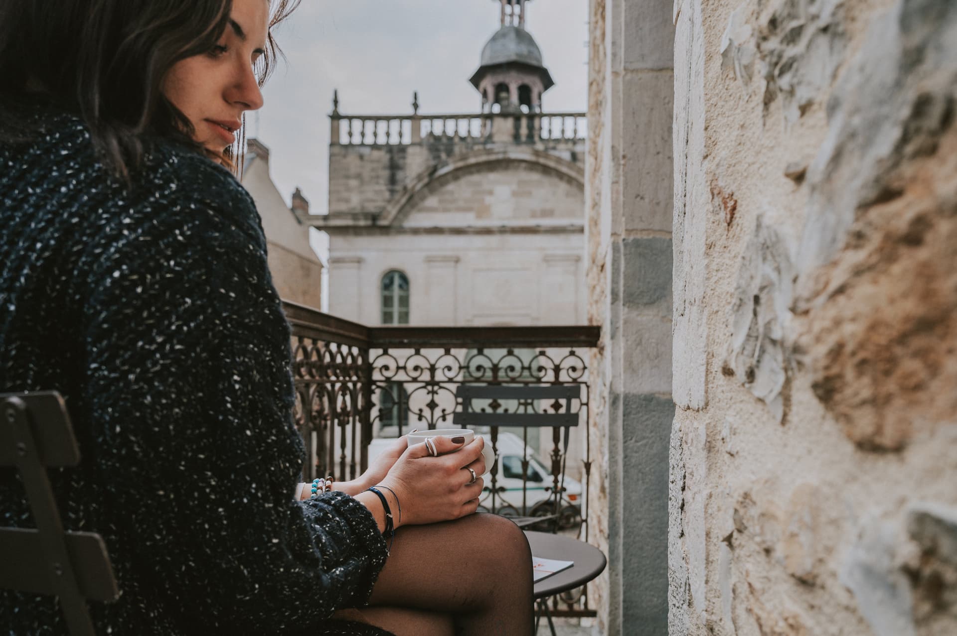 Une femme se réchauffe les mains avec une tasse de café sur le balcon de sa chambre à l'Hôtel Les Fleurines, avec une vue magnifique sur la Chapelle des Pénitents Noirs à Villefranche-de-Rouergue, Aveyron.