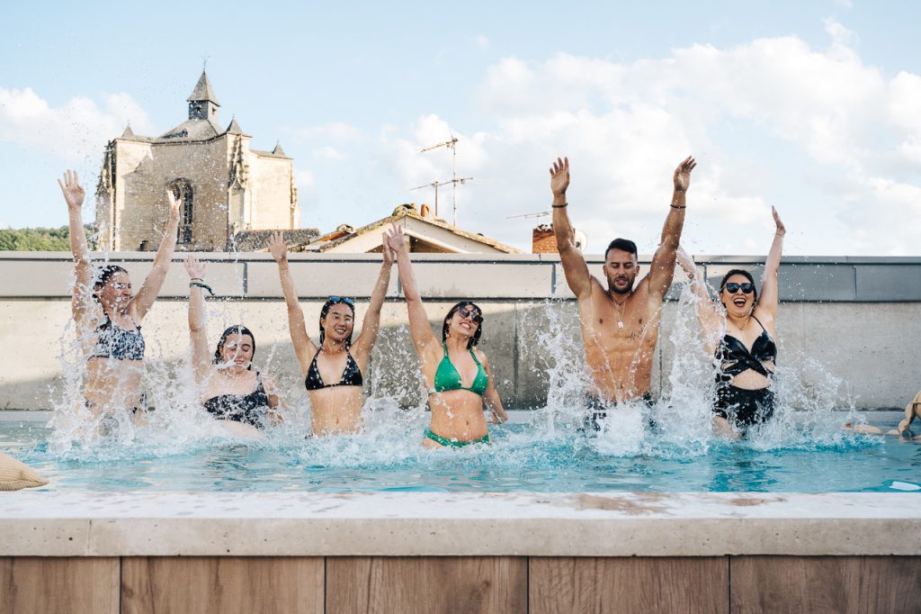 Un homme et des femmes sautent dans la piscine en rooftop de l’Hôtel Les Fleurines à Villefranche-de-Rouergue, Aveyron, avec une vue spectaculaire sur la Collégiale Notre-Dame. L’atmosphère est dynamique et estivale, parfaite pour profiter d’un moment de détente et d’amusement sous le soleil.