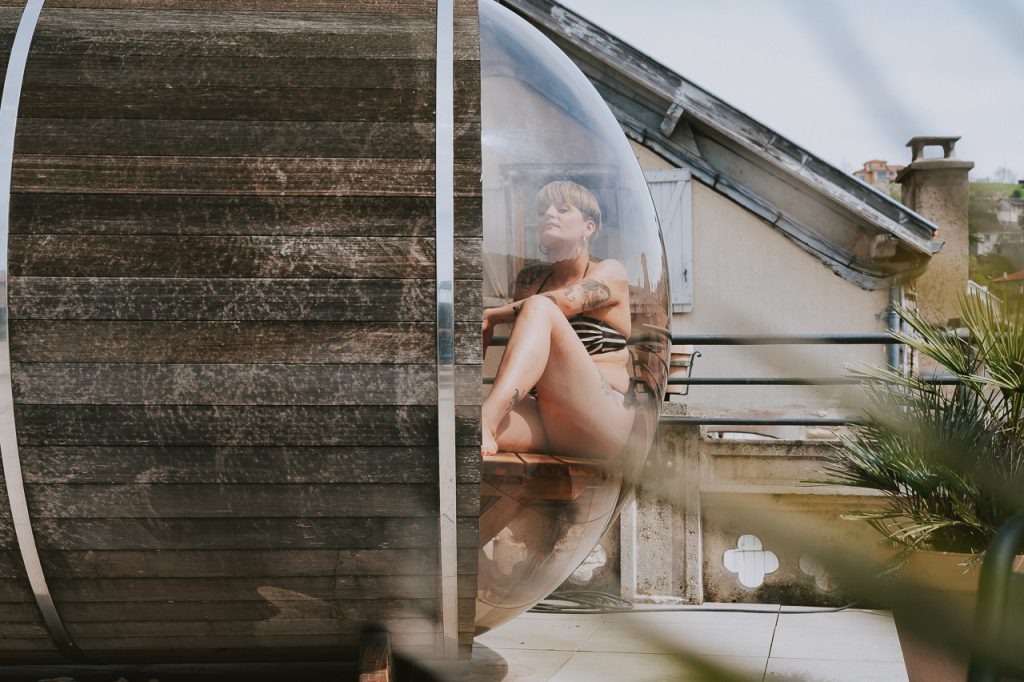 Une femme profite pleinement d'un instant de relaxation dans le sauna panoramique de l'Hôtel Les Fleurines à Villefranche-de-Rouergue, en Aveyron, avec une vue imprenable sur les toits de la bastide. Un moment de bien-être unique, au cœur de l'Aveyron, pour se ressourcer en pleine nature.