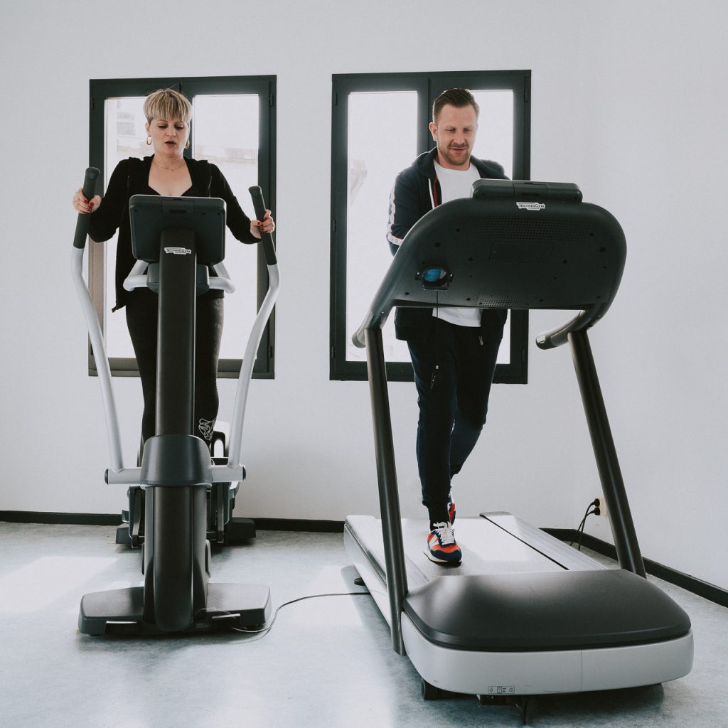 Un couple fait du sport dans la salle de sport moderne de l'Hôtel Les Fleurines à Villefranche-de-Rouergue, Aveyron. Un lieu parfait pour combiner bien-être et activité physique lors de votre séjour.