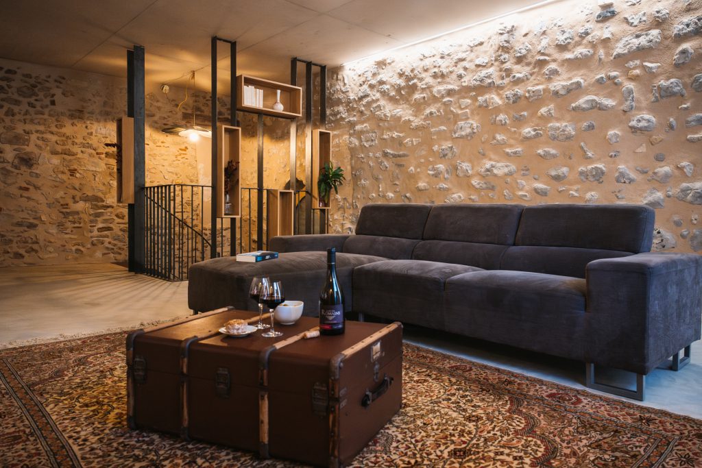 Salon de la Maison des Cassiers, au sein de l’Hôtel Les Fleurines à Villefranche-de-Rouergue, Aveyron. Son mur en pierre apparente, son sol en béton ciré, son tapis à motifs et ses étagères en bois et métal créent une atmosphère à la fois authentique et contemporaine, idéale pour un moment de détente.