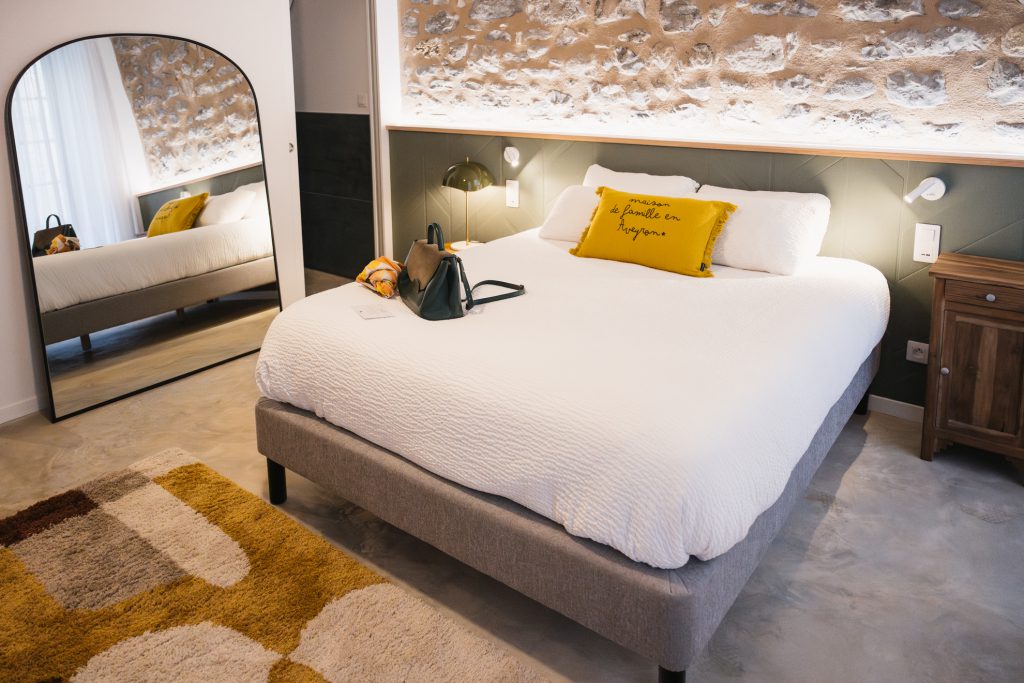 Chambre au charme authentique de la Maison des Cassiers, située dans l’Hôtel Les Fleurines à Villefranche-de-Rouergue, Aveyron. Son mur en pierre, sa tête de lit en tapisserie verte texturée, son grand miroir et ses coussins lumineux créent une atmosphère chaleureuse et élégante, parfaite pour un séjour tout en confort.