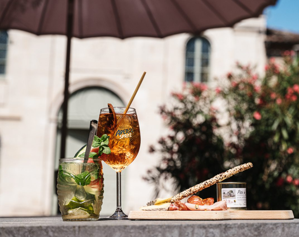 Apéritif convivial avec spritz, mojito et planche de charcuterie sur la terrasse de l’Hôtel Les Fleurines à Villefranche-de-Rouergue, offrant une vue sur la chapelle des Pénitents Noirs et la place Saint-Jacques, au cœur de l’Aveyron.