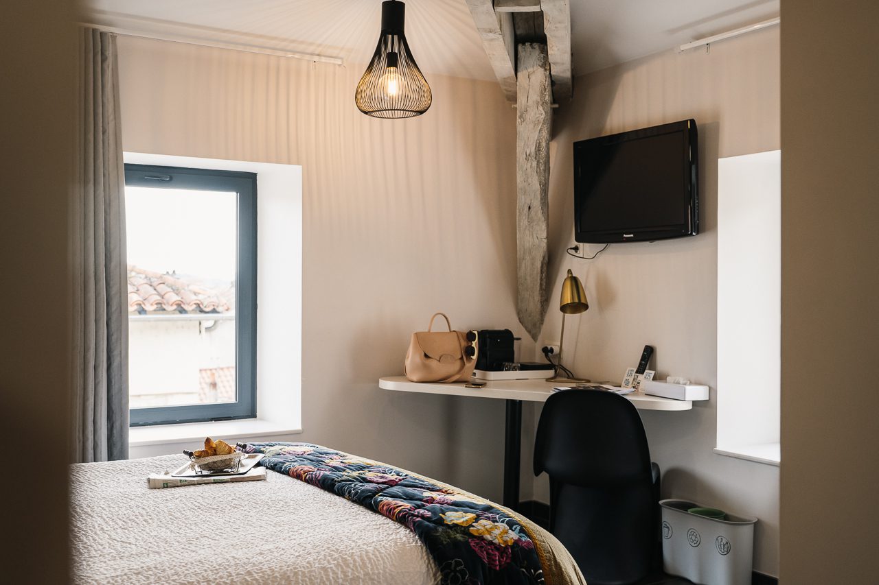 Chambre spacieuse et lumineuse de l'Hôtel Les Fleurines à Villefranche-de-Rouergue, avec des poutres apparentes, alliant authenticité et modernité. Parfaite pour se détendre ou travailler, offrant une vue imprenable sur les toits de la ville.