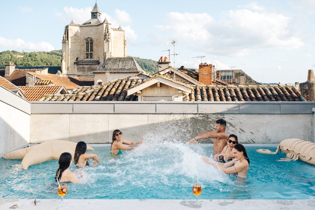 Des personnes profitent de la piscine en rooftop de l’Hôtel Les Fleurines à Villefranche-de-Rouergue, Aveyron, offrant une vue imprenable sur la Collégiale Notre-Dame et les toits de la bastide. Un moment de détente unique dans un cadre d’exception.