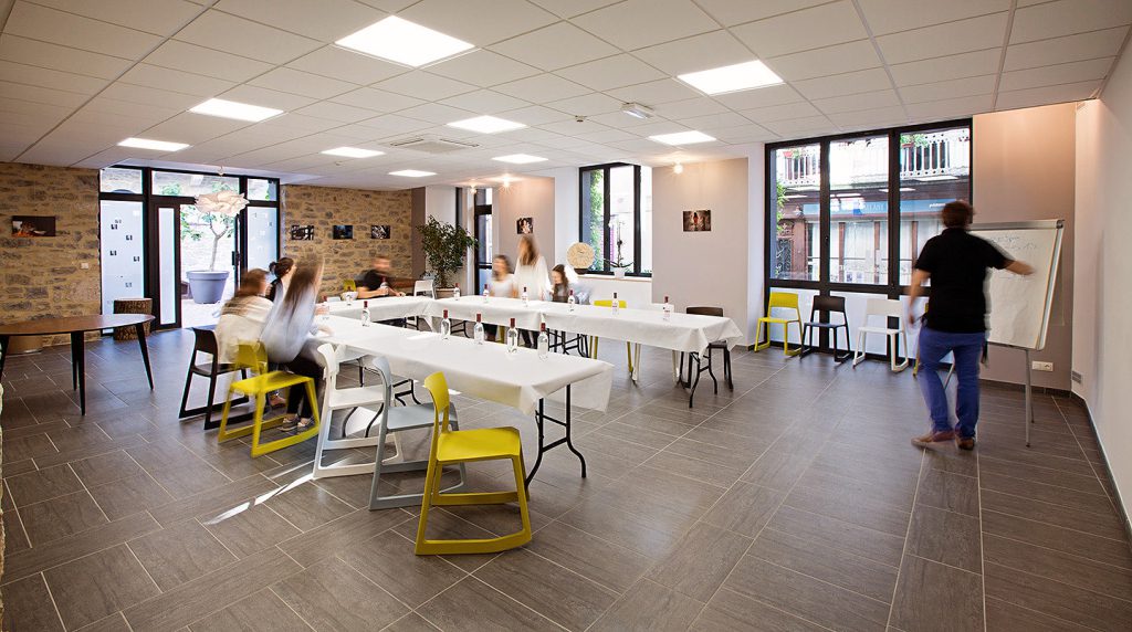 Salle de séminaire de l’Hôtel Les Fleurines à Villefranche-de-Rouergue, Aveyron, aménagée avec des tables blanches et des chaises colorées. Des participants sont en plein séminaire, dans un espace lumineux et moderne, avec un mur en pierre et blanc qui crée une ambiance à la fois professionnelle et accueillante.