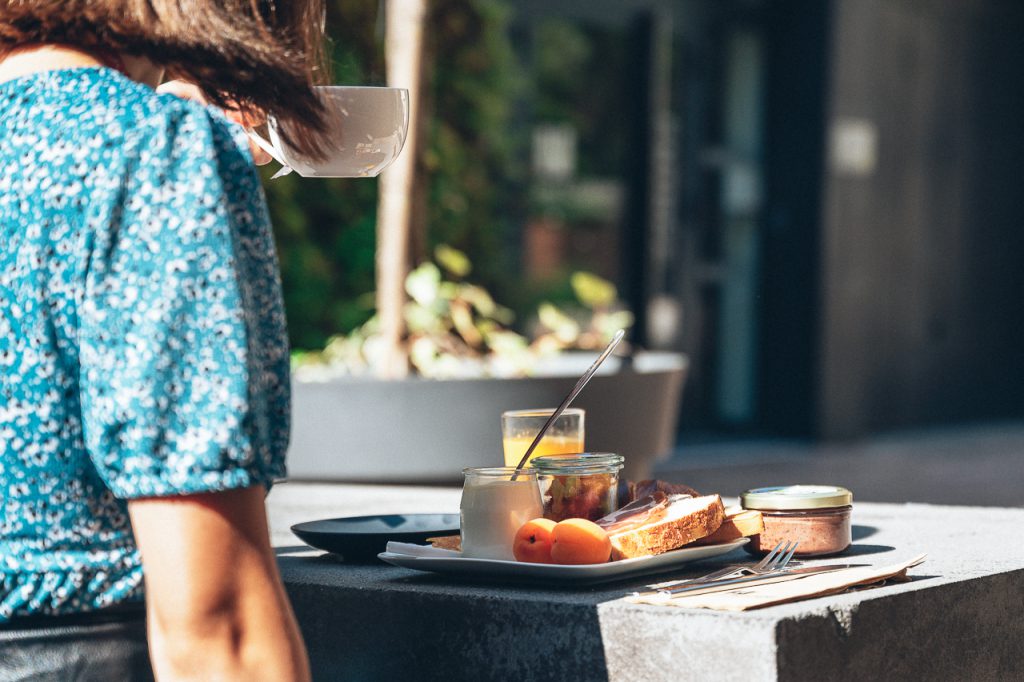 Petit-déjeuner en terrasse à l'Hôtel Les Fleurines à Villefranche-de-Rouergue, Aveyron, avec des produits locaux et de saison : pain artisanal, jambon du terroir, yaourt fermier, fruits frais, confiture maison et jus d'orange. Une expérience gourmande et écoresponsable dans un cadre verdoyant, face au mur végétal et la Chapelle des pénitents noirs.